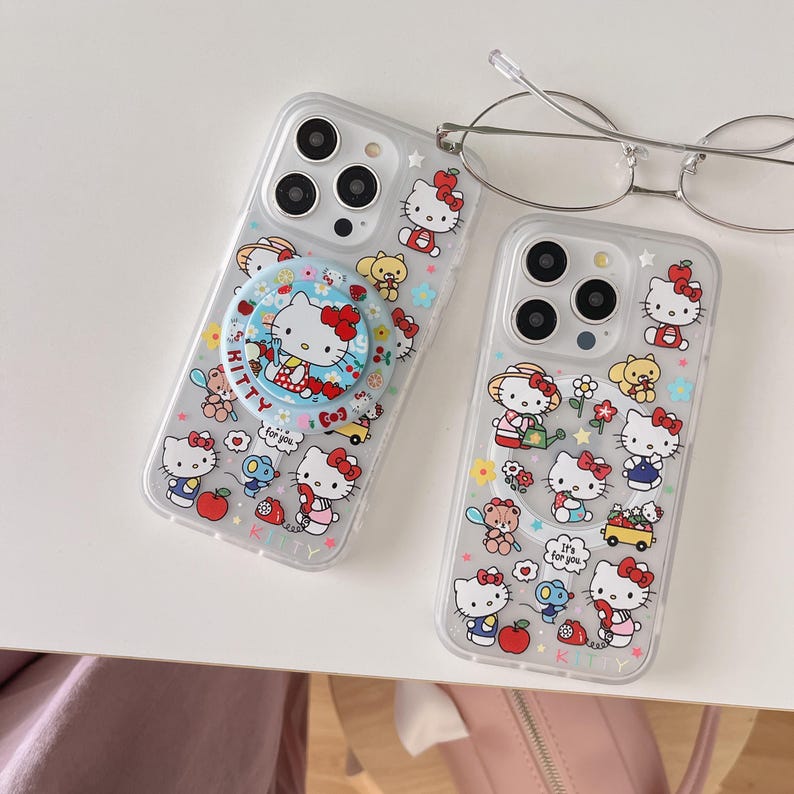 May include: Two clear phone cases with Hello Kitty designs. One case has a pop socket with a Hello Kitty graphic and the text "Kitty". The other case features various Hello Kitty illustrations with flowers and other characters.