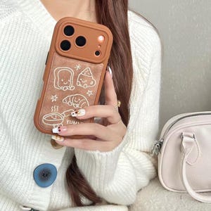May include: A brown phone case with a textured panel. It features white line drawings of food items, including bread, a croissant, and a chef's hat. The case has a protective frame and a camera cutout.