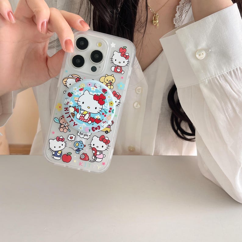 May include: Clear phone case featuring a Hello Kitty design with a pop-up grip. The case has a variety of Hello Kitty illustrations, including the character with a red bow, flowers, and other cute elements. The pop-up grip has the words "Hello Kitty" on it.