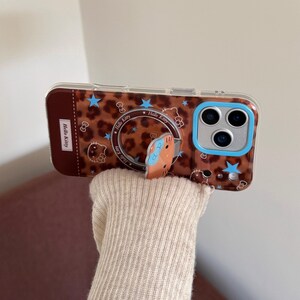 以下が含まれることがあります： ヒョウ柄のデザインとハローキティのデザインが施された、茶色とベージュの携帯電話ケース。ケースには青いカメラの縁と、ハローキティのキャラクターが付いたポップアップ式の電話グリップが付いています。ケースは人が持っています。
