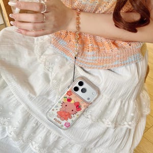 May include: Clear phone case with a cartoon character design, featuring a pink bear with a bow and floral accents. The case is attached to a gold-toned beaded wrist strap. The phone case is on a white skirt.