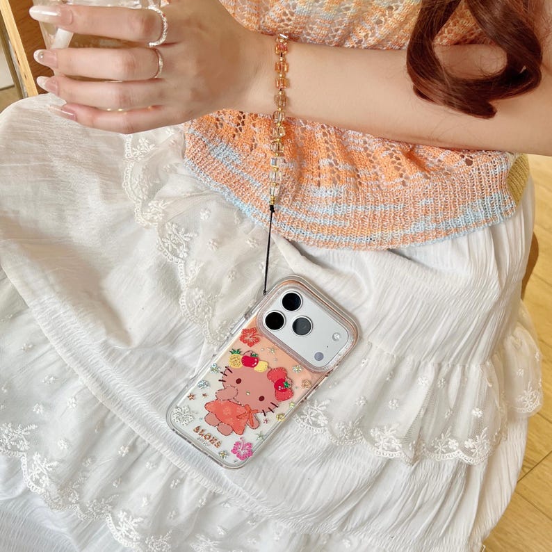 May include: Clear phone case with a cartoon character design, featuring a pink bear with a bow and floral accents. The case is attached to a gold-toned beaded wrist strap. The phone case is on a white skirt.