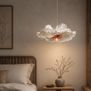 Puede incluir: Una lámpara colgante blanca con forma de flor cuelga sobre una cama. La lámpara tiene un diseño con volantes y un centro rosa. El dormitorio tiene una paleta de colores neutros con una estructura de cama de madera, una mesita de noche y una impresión botánica enmarcada.