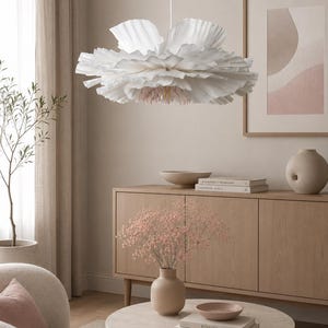 Peut inclure: Une suspension lumineuse blanche en forme de grande fleur à plusieurs couches avec des pétales froncés. Le centre de la lampe présente de fins filaments rose clair suspendus vers le bas, fixée au plafond par un cordon blanc fin.