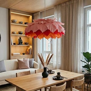 Può includere: Una grande lampada a sospensione a strati nei toni del rosa, del rosso e dell'arancione è appesa sopra un tavolo da pranzo in legno. La lampada assomiglia a un fiore. La stanza presenta una palette di colori neutri con un divano, mensole e una pianta in vaso.