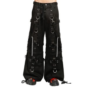 Puede incluir: Pantalones negros de pierna ancha con múltiples correas verticales de tela, anillos en D de metal plateado y remaches. La prenda incluye dos cadenas de metal plateado colgantes unidas a las correas y un cierre de cordón en la cintura.
