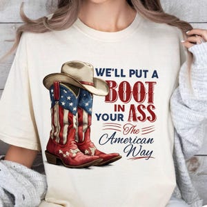 May include: Cream-colored t-shirt featuring a pair of red, white, and blue cowboy boots and a cowboy hat. The boots are decorated with the American flag. Text on the shirt reads "We'll put a boot in your ass the American way."