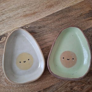 Op de afbeelding: Twee keramische avocado-vormige schaaltjes. Eén is gebroken wit met een gele cirkel en een lachend gezicht. De andere is groen met een bruine cirkel en een lachend gezicht. De schaaltjes staan op een houten oppervlak.