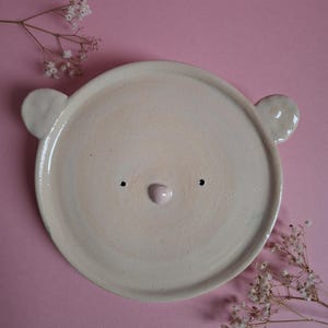 Op de afbeelding: Een crèmekleurige keramische plaat in de vorm van een berengezicht, met twee kleine oren, twee zwarte stippen voor ogen en een roze neus. Op een roze achtergrond, met takjes witte bloemen.