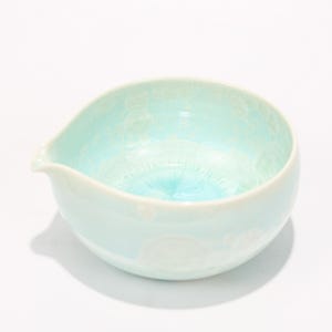 May include: A light blue ceramic bowl with a spout. The bowl has a smooth, glossy finish and a unique crystalline pattern on the interior. The bowl is designed with a rounded shape and a small spout for pouring.