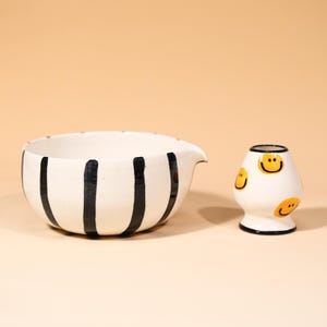 May include: A white ceramic bowl with black vertical stripes and a spout, next to a small white ceramic vessel decorated with yellow smiley faces. Both items are on a light beige surface.