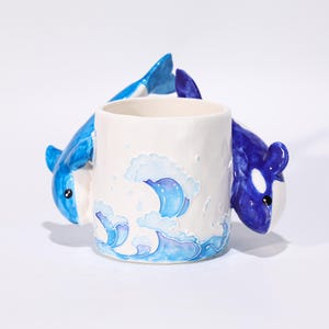 Może przedstawiać: Biały ceramiczny kubek z wzorem niebieskich fal oceanu i dwiema figurkami orek. Orki są niebiesko-białe, po jednej z każdej strony kubka. Kubek ma błyszczące wykończenie.