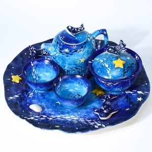Può includere: Un servizio da tè in ceramica blu con un tema celeste. Il set include una teiera, una ciotola con coperchio, due tazze e un vassoio, tutti decorati con stelle bianche e motivi a spirale. Accenti a forma di stella gialla e una statuina a forma di balena completano il set.