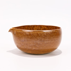 Puede incluir: Un cuenco de cerámica marrón con un pico vertedor. El cuenco tiene forma redondeada y superficie texturizada. El color es marrón cálido y terroso, con variaciones de tono. El pico es pequeño y curvado.