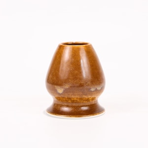 May include: A brown ceramic matcha bowl with a rounded body and a wide base. The bowl has a glazed finish with variations in color, ranging from light to dark brown. The interior is a darker shade of brown.