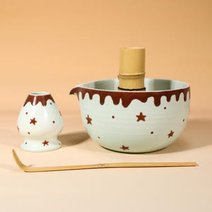 May include: A light green ceramic matcha bowl with a brown drip pattern and brown star accents, a matching ceramic whisk holder, and a bamboo whisk. A long, thin, curved bamboo tea scoop lies horizontally in front of the bowl and holder.