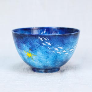 Könnte beinhalten: Eine blaue Keramikschale mit einer gemalten Meereslandschaft. Die Schale hat einen Farbverlauf von Blautönen, mit weißen Pinselstrichen, die Fischen ähneln, und einem gelben Stern. Die Schale hat eine runde Form und steht auf einem kleinen Sockel.
