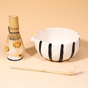 May include: A matcha tea set featuring a white ceramic bowl with black stripes, a bamboo whisk, and a bamboo scoop. The whisk has a white base with yellow smiley faces. The set is designed for preparing matcha tea.