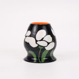 May include: A black ceramic vase with white floral designs and green leaves. The interior of the vase is orange. The vase has a rounded body and a flared base. The floral design is stylized and modern.
