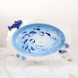 May include: An oval ceramic serving platter with a white scalloped edge and a blue beaded border. The center features a painted scene of blue fish swimming in light blue water. Three-dimensional ceramic sea creatures adorn the rim.