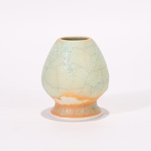 May include: A ceramic matcha bowl with a light green and orange glaze. The bowl has a rounded body and a flared base. The surface has a crackle pattern. The bowl is designed for whisking matcha tea.