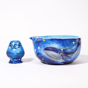 May include: A blue ceramic matcha bowl and whisk holder set. The bowl features a whale design with stars and a celestial background. The whisk holder is a small, blue, vase-shaped container with a painted design. The set is designed for preparing matcha tea.