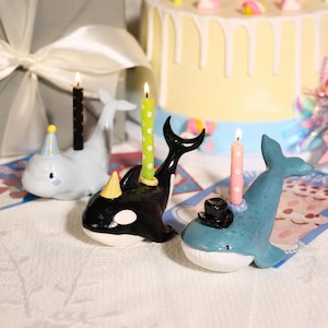 May include: Three whale-shaped candle holders made of ceramic. One light blue whale wears a yellow party hat with a black polka-dot candle. One black and white orca wears a yellow party hat with a green polka-dot candle. One blue whale wears a black top hat with a pink polka-dot candle.