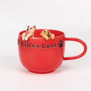 Puede incluir: Una taza de cerámica roja con asa, con dos pequeñas figuras de perros asomándose por el borde. La taza está inscrita con "Bella & Luna" e incluye un diseño de huella de pata. La taza es de color rojo vibrante.