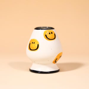 May include: A white ceramic matcha whisk holder with a black rim and base. The holder is decorated with yellow smiley faces. The product is designed to hold a matcha whisk, keeping it in good shape.