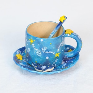 May include: A ceramic teacup and saucer set with a matching spoon. The set is painted in shades of blue with white swirls and yellow stars. A small whale figurine is on the saucer. The handle of the cup and the spoon are decorated with yellow stars.