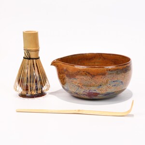 Puede incluir: Un juego de té matcha con un cuenco de cerámica con boquilla, un batidor de bambú y una cuchara de bambú. El cuenco tiene un esmalte marrón y azul. El batidor y la cuchara son de bambú.