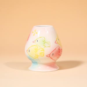 Könnte beinhalten: Eine weiße Keramikvase mit rundem Körper und ausgestelltem Fuß. Die Vase ist mit farbenfrohen, skurrilen Zeichnungen von Vögeln in Gelb, Grün und Rot verziert. Der Sockel hat einen Farbverlauf aus Blau, Rosa und Gelb.