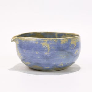 Puede incluir: Un cuenco de cerámica hecho a mano con un pico vertedor. El cuenco presenta un esmalte azul con un aspecto moteado y texturizado y un borde de color beige claro. El cuenco es redondo y se apoya sobre una superficie blanca.