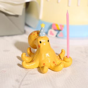 May include: A yellow octopus figurine with orange spots, wearing a blue party hat with a yellow star on top. The octopus has a smooth, glossy finish and features a small hole in one of its tentacles that holds a thin, light pink birthday candle.