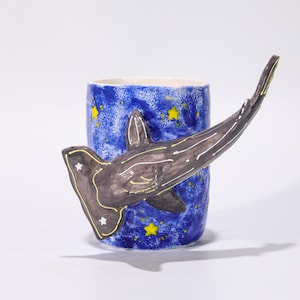 May include: A ceramic mug with a blue background and yellow star pattern. A hammerhead shark handle is attached to the mug. The shark is dark grey with white and yellow accents. The mug is on a white background.