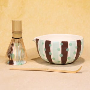 Può includere: Un set da tè matcha con una frusta di bambù, una ciotola di ceramica con strisce marroni e pois verdi e un cucchiaio di bambù. La ciotola ha un beccuccio. La frusta ha un manico marrone chiaro.