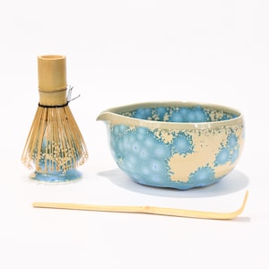 May include: A matcha tea set comprising a light blue and cream ceramic bowl, a bamboo whisk, and a bamboo scoop. The bowl has a distinctive textured pattern. The whisk is made of bamboo and is tied with black string.