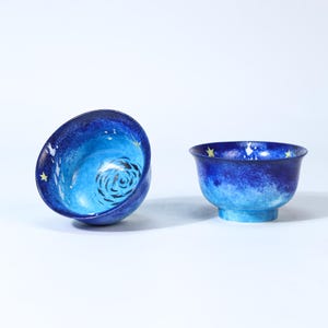 May include: Two blue ceramic bowls with a celestial design. One bowl is tilted, revealing an interior swirl pattern. Both bowls feature a gradient of blue hues, with yellow star accents. The bowls are set against a white background.
