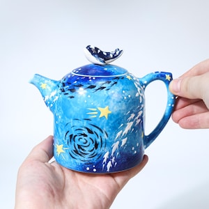 Può includere: Una teiera in ceramica blu con un design a tema marino. La teiera presenta una maniglia del coperchio a forma di balena ed è decorata con pesci, stelle e motivi a vortice. Il manico e il beccuccio sono anch'essi blu, con accenti di stelle gialle.