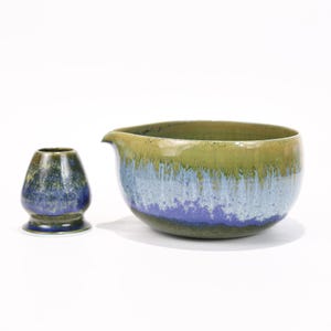 Könnte beinhalten: Ein Keramik-Matcha-Schalen- und Schneebesenhalter-Set. Die Schale hat einen Ausguss und ist in Grün-, Blau- und Lilatönen glasiert. Der Schneebesenhalter ist ein kleines, abgerundetes Gefäß mit einer ähnlichen blau-grünen Glasur.