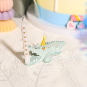 以下が含まれることがあります： 小さな黄色いパーティーハットをかぶったライトグリーンのワニのフィギュア。口を開けて、小さな金色の星模様が描かれた細い白いバースデーキャンドルをくわえています。ワニには目に見える歯とオレンジ色の目があります。