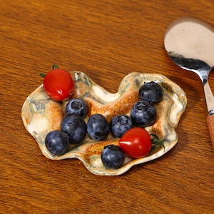 Puede incluir: Un pequeño plato de cerámica de forma única, con una paleta de colores terrosos moteados, que contiene arándanos frescos y tomates cherry rojos. Una cuchara de acero inoxidable con mango de madera descansa cerca, sugiriendo servir o preparar.
