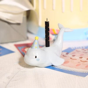 May include: A light blue beluga whale figurine wearing a small blue party hat with yellow polka dots. The whale has a black candle with gold star patterns inserted into its back. The figurine features simple black eyes and a small blue cheek mark.