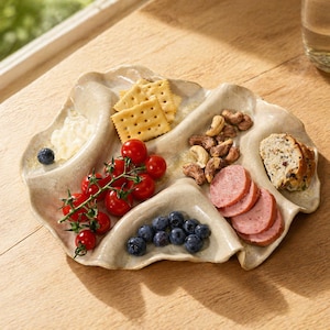 May include: A beige, multi-compartment serving platter filled with snacks. It holds crackers, cherry tomatoes, blueberries, cashews, sliced sausage, and bread. A small dollop of white spread and a single blueberry are also present.