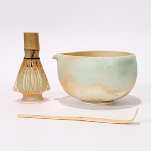 Puede incluir: Un juego de té matcha con un cuenco de cerámica, un batidor de bambú y una cuchara de bambú. El cuenco es de color verde claro y marrón con un pico. El batidor y la cuchara son de bambú.