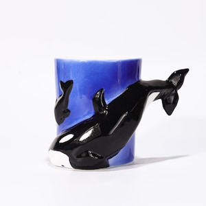 May include: A ceramic mug featuring a black and white orca whale design. The mug has a blue interior and exterior, with the whale's body forming the handle. The orca is depicted with its distinctive markings.