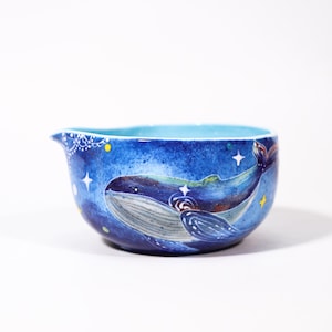 May include: A ceramic bowl with a blue interior and exterior. The exterior features a hand-painted whale design with stars and a celestial background. The bowl has a spout and a decorative white lace pattern near the rim.