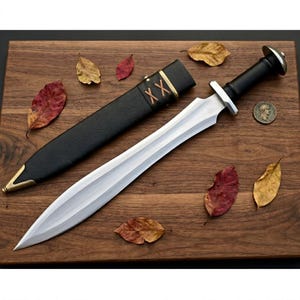 Könnte beinhalten: Ein silbernes und schwarzes Schwert mit einer Lederscheide. Das Schwert hat eine zweischneidige Klinge und einen schwarzen Griff. Die Scheide ist schwarz mit goldenen Akzenten und braunen Nähten. Das Schwert und die Scheide werden auf einer Holzoberfläche mit Herbstblättern präsentiert.