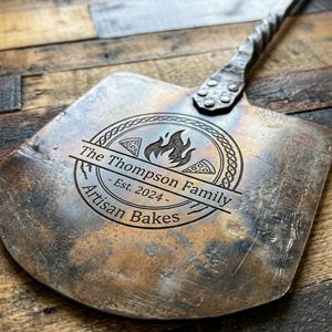 Könnte beinhalten: Ein Pizzaschieber aus Metall mit einem langen, gedrehten Griff und einer flachen, breiten Schaufel. Die Oberfläche zeigt ein eingraviertes kreisförmiges Logo mit einem Flammensymbol, zwei Pizzastücken und dem Text: The Thompson Family - Est. 2024 - Artisan Bakes.