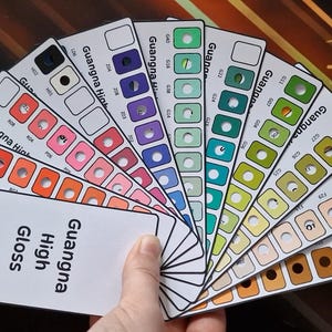 Puede incluir: Una mano sostiene un abanico de muestras de color etiquetadas como "Guangna High Gloss". Cada muestra muestra una serie de muestras de color en varios tonos, incluyendo rojo, rosa, morado, verde y marrón. El número "168" es visible.